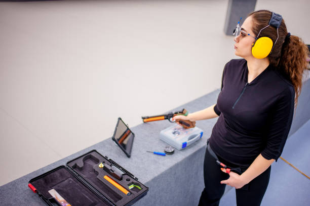 A determined female athlete aiming a precision air pistol during practice at an indoor shooting range. She wears protective red earmuffs and specialized shooting glasses, highlighting her focus and professionalism. The background features a modern shooting range setup with digital scoring tablets and shooting equipment. This image captures the essence of competitive shooting sports, concentration, and modern technology in athletics.