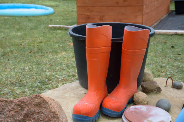 A pair of bright orange rubber boots placed on a stone surface near a black bucket, with a backyard and wooden structure in the background