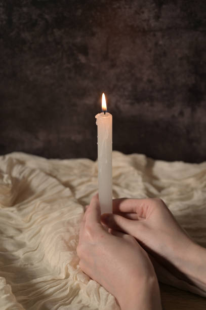 istock One white lit candle in woman hands on dark gray background, beige cloth 2189249321