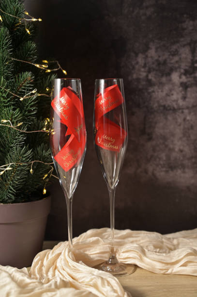istock Christmas decor, two transparent glass with a red ribbon inside, tree with lights, dark background, beige cloth on wooden table 2189249324