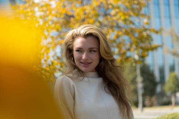Portrait of beautiful smiling serene long haired blond caucasian woman in white sweater standing outdoors on city street in a sunny autumn day by tree with yellow leaves. Soft focus. People theme.
