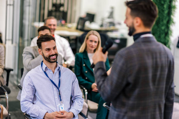 istock Young business man lecturing his multiethnic colleagues. Having a speech while holding a microphone. Everyone is listening and some are raising their hand. 2189252617