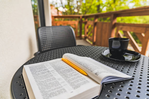 istock Open Holy Bible with yellow pen lying on plastic garden table with black coffee cup standing on balcony with wooden fence and green surroundings 2189259891