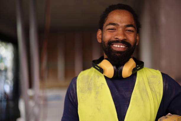 A confident construction worker smiles warmly, wearing a high-visibility vest and yellow headphones at a bustling job site. His positive attitude reflects dedication and professionalism in the construction industry.