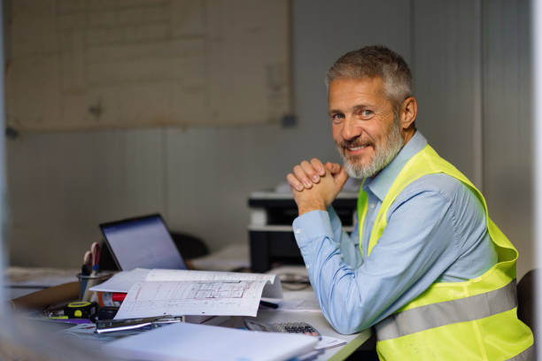 Confident Construction Worker Smiling at Desk with Blueprints and Laptop