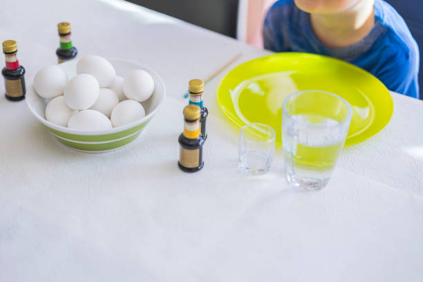 Child at table during Easter egg preparation, surrounded by food coloring bottles and white eggs in bowl. Sweden.