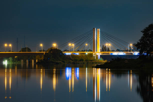 Wroclaw, Poland - May 11 2024: Beautiful nightscape of high trees and bushes with Millenium bridge between and reflecting in silent Odra river at cloudy and starry night