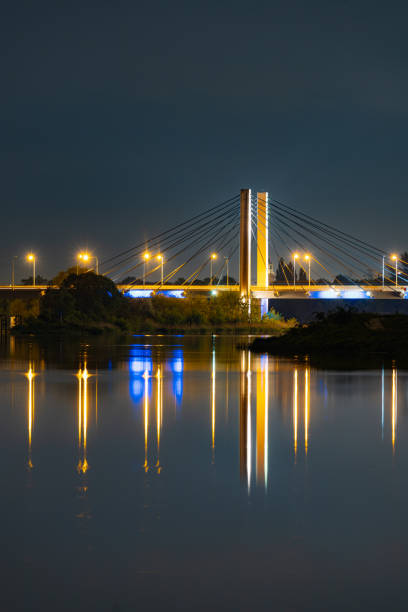 Wroclaw, Poland - May 11 2024: Beautiful nightscape of high trees and bushes with Millenium bridge between and reflecting in silent Odra river at cloudy and starry night