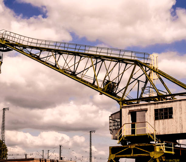 A crane is seen in the sky above a building. The crane is old and rusty, and the sky is cloudy. Scene is somewhat somber and nostalgic, as the crane seems to be a relic of a bygone era