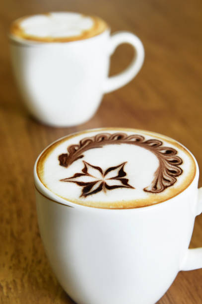 A white cup with colorful, circular patterns holds coffee on a grassy surface. Next to it, a vivid yellow notebook is adorned with a white flower, possibly a daisy. A lace-bordered fabric adds a delicate backdrop, creating a charming, serene setting, suggesting a casual, outdoor break or planning session.