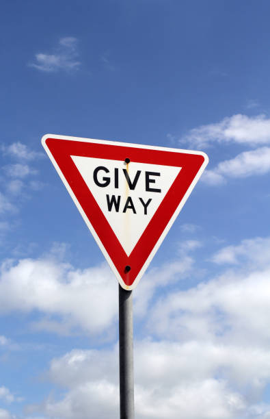 A white triangle road sign with a red border is mounted on a metal pole, set against a blurred background of trees and grass. Sign 2.