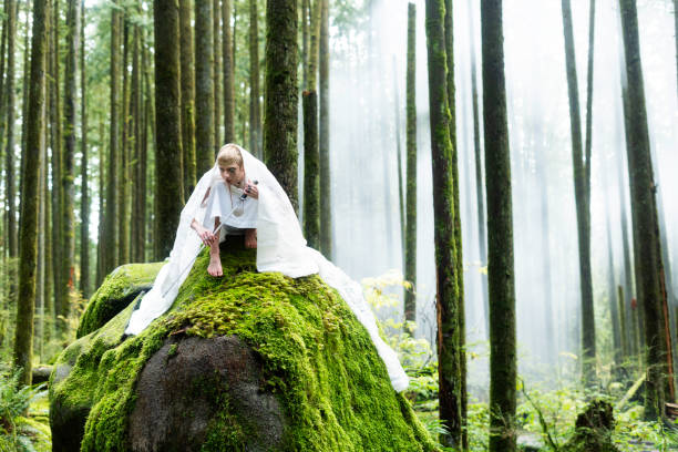 Elfin male in a long white cape fantasy costume poses on a huge mossy rock in Golden Ears Provincal Park, Vancouver, British Columbia, Canada