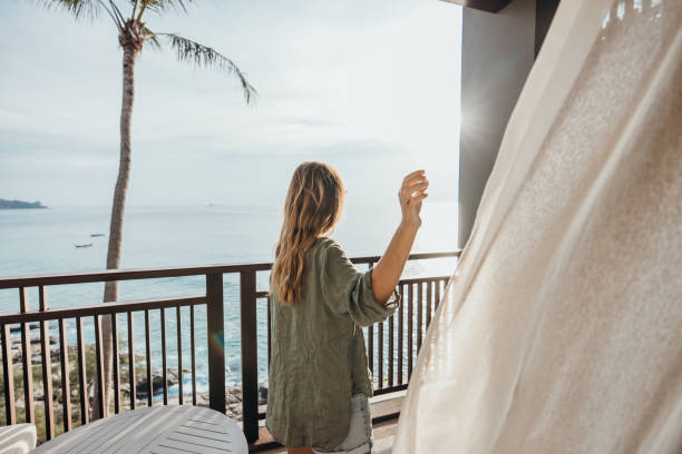 Woman in casual attire stands on a wooden balcony, gazing at the ocean. She holds a curtain aside, revealing a serene view of the sea and palm trees in the distance.