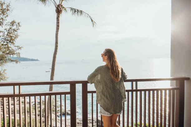 A woman leans on a wooden balcony, gazing at the ocean during sunset. Palm trees frame the serene view, capturing a moment of relaxation and tranquility.