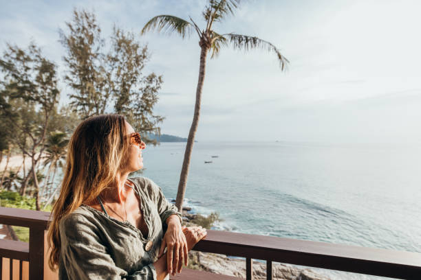 A woman leans on a wooden balcony, gazing at the ocean during sunset. Palm trees frame the serene view, capturing a moment of relaxation and tranquility.