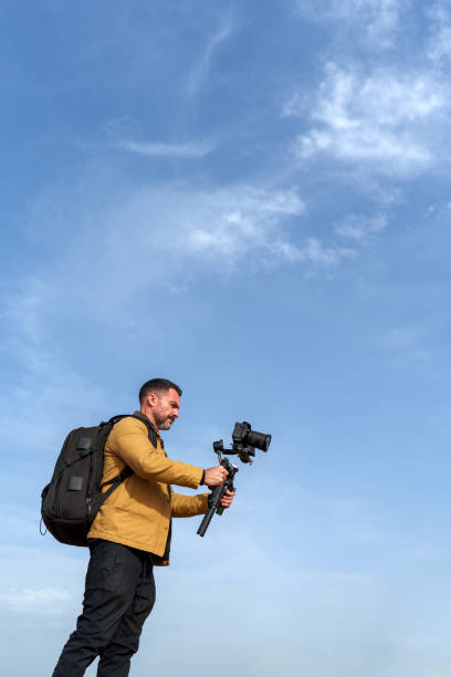 istock Videographer using a gimbal and camera to film outdoors against a vivid blue sky and fluffy white clouds 2189335129