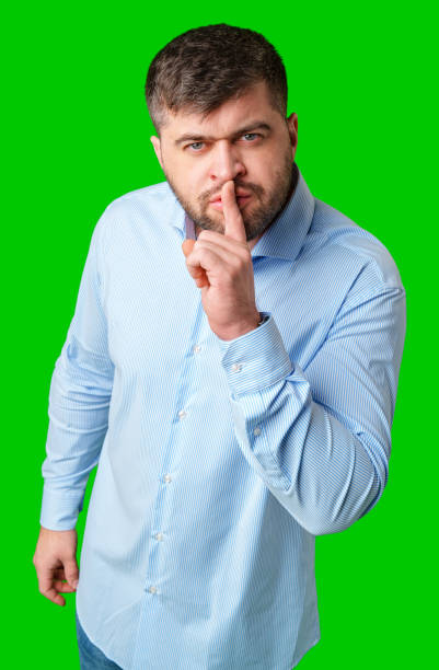 istock A man in a blue shirt gestures for silence with a finger on his lips against a bright green background 2189339307
