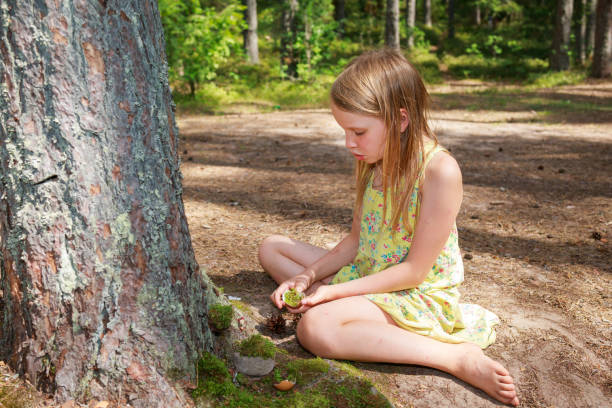 A young girl in a yellow floral dress sits in a sunlit forest, creating a miniature scene at the base of a moss-covered tree trunk