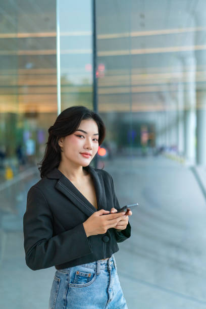 istock Asian Chinese Woman Looking at Camera while Using Phone Near Large Glass Windows with Urban Reflections 2189349152