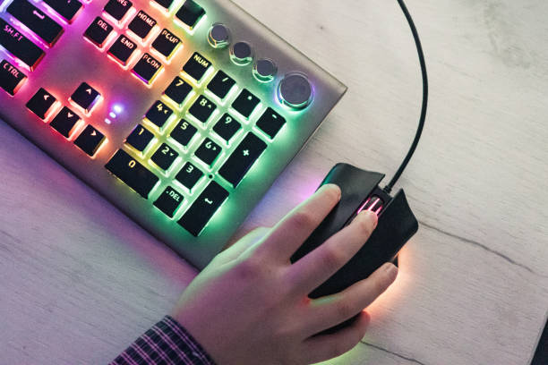 A close-up view of a wireless computer mouse placed on a black mouse pad next to a closed laptop on a wooden desk. Warm decorative lights create a soft bokeh in the background, adding a cozy contrast to the dark tech setup.