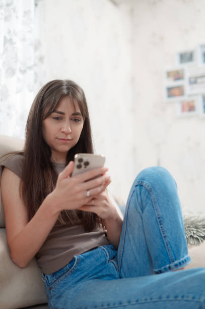 istock Young happy woman sitting comfortably on sofa, smiling while using her phone indoors during a relaxing afternoon vertical 2189370740
