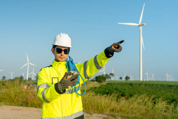 istock Wind turbine or windmill engineer worker hold and look to mobile phone also point to his left side with row of wind turbine in the background. 2189377427