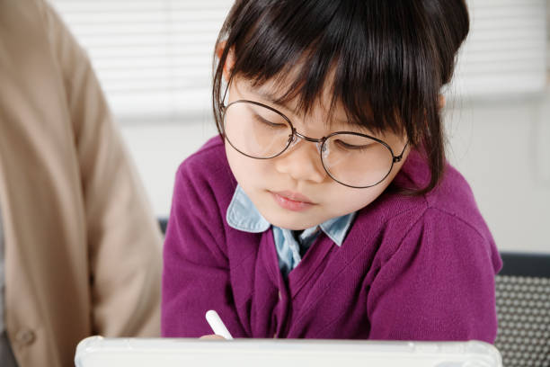 An elementary school girl with glasses studying while operating a tablet. She is receiving tutoring.