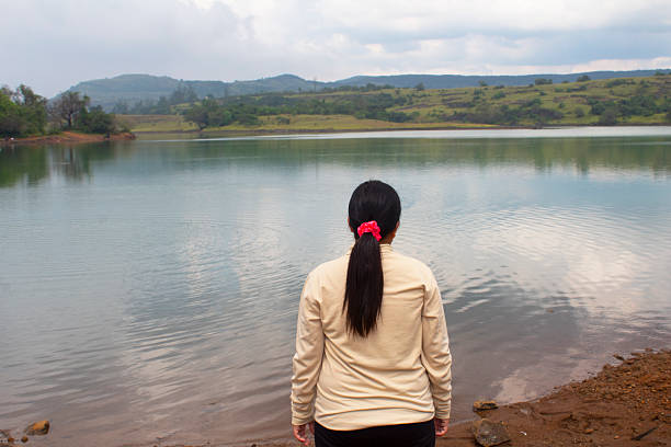 Female Hiker Stands on the Shore of a Picturesque Lake, Gazing Out at the Calm Waters Beneath a Cloudy Sky. The Scene Exudes Serenity, Highlighting the Deep Connection of Hiker with Nature. This Moment Captures the Essence of Adventure, Self-Reflection, and the Tranquility of Outdoor Exploration