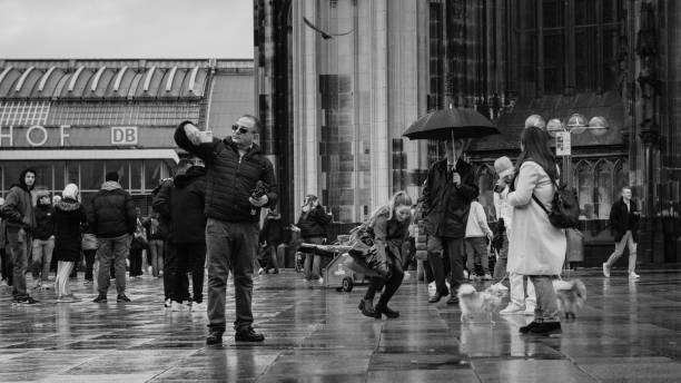 Cologne, Germany - October 27, 2023: In front of Cologne Cathedral, tourists brace the rain under umbrellas. A man throws food for birds while others watch, adding life to the wet, reflective cobblestones. The mix of modern and historical charm reflects a serene yet lively moment.