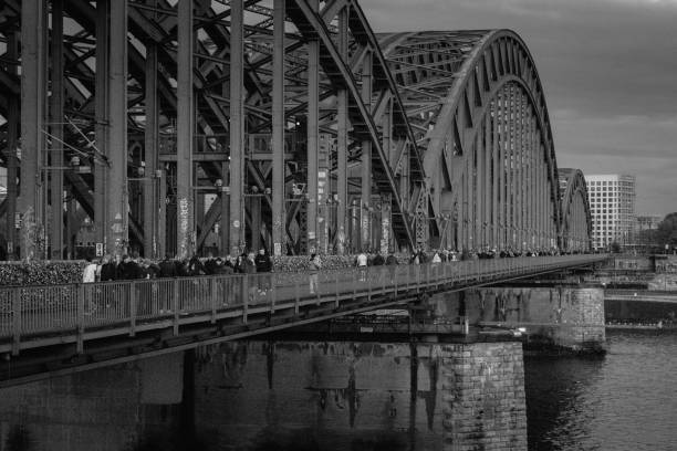 Cologne, Germany - October 27, 2023: The intricate steel beams of the Hohenzollern Bridge create a powerful urban backdrop, contrasting with countless love locks adorning the railings. Visitors walk across the bridge, adding motion and life to this industrial yet romantic scene.