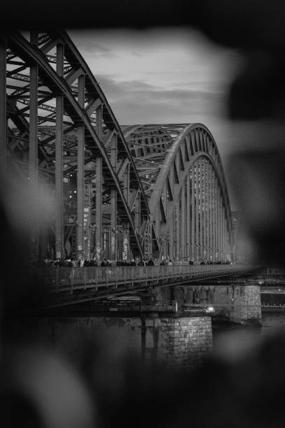 Cologne, Germany - October 27, 2023: The Hohenzollern Bridge is captured in the soft light of twilight, its robust steel structure dominating the frame. The evening air brings a calm mood, while people leisurely stroll across the iconic bridge. Reflections on the river below enhance the autumn atmosphere.