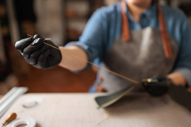 Craftswoman is seen sewing the edges of a leather wallet using a needle and thread in her well-equipped workshop.