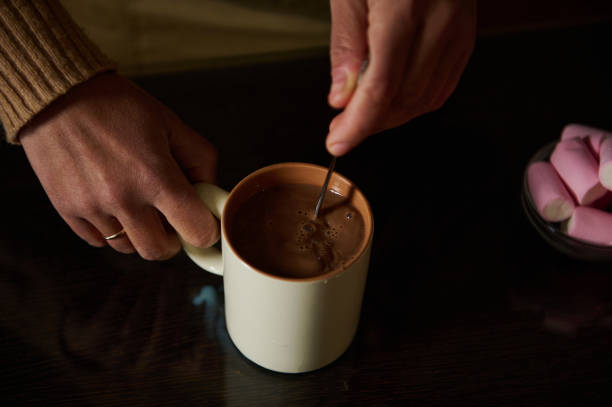 Hands shown stirring a cup of hot chocolate on a table beside marshmallows.