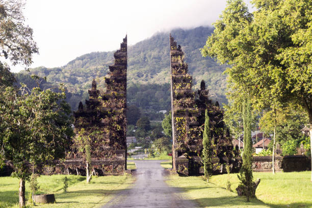 This is a color photograph of traditional Indonesian gates opening up to a road surrounded with green tropical plants in Bali, Indonesia.