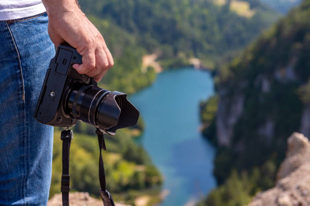 Close-up of photographer's hand with camera while standing on cliff overlooking lake during travel.