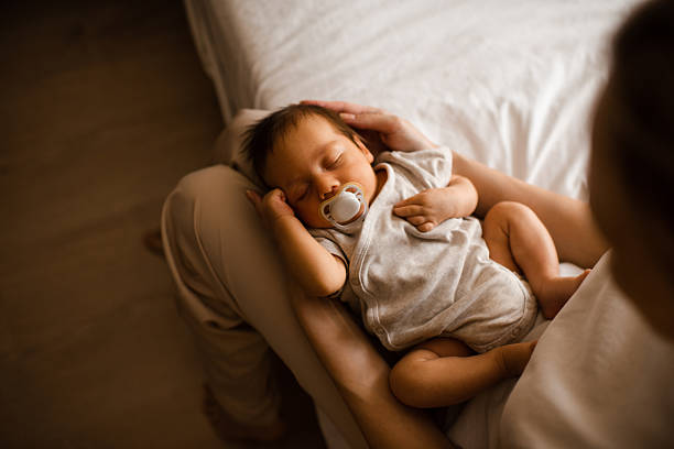 Sleeping baby boy 1-2 months old with pacifier sleeping on mother hands in bedroom closeup top view. Motherhood.