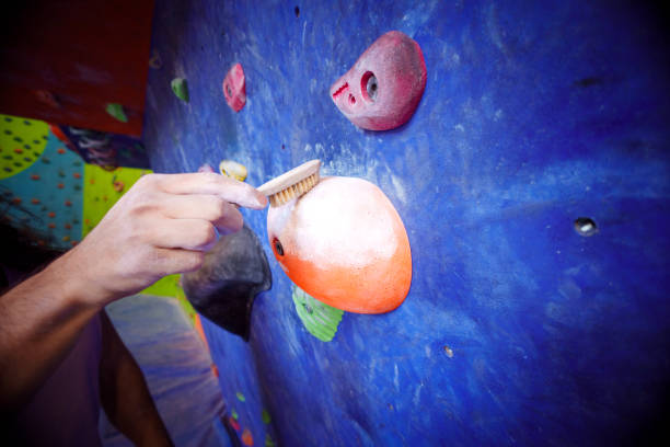 A close-up of a climber using a brush to clean a handhold on an indoor climbing wall, ensuring optimal grip and safety. The scene highlights the attention to detail and preparation required in climbing. The vibrant background features colorful holds and sections of the climbing gym, emphasizing the dynamic environment of the sport. This image showcases the importance of maintenance and technique in rock climbing activities.
