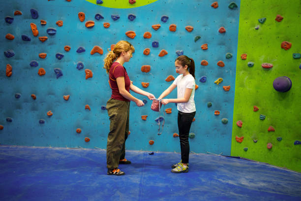 Two climbers preparing for indoor rock climbing by applying chalk to their hands. The background features a vibrant blue climbing wall with colorful holds, emphasizing the indoor climbing environment. The image captures the importance of teamwork and preparation in rock climbing
