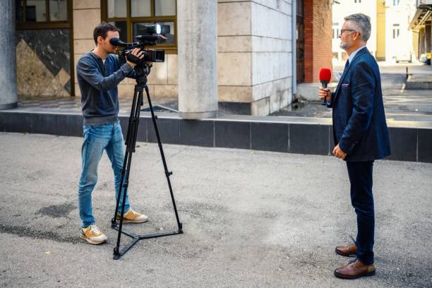 A male television reporter holding a microphone, is standing outdoors in front of a building. A male camera operator is filming him.