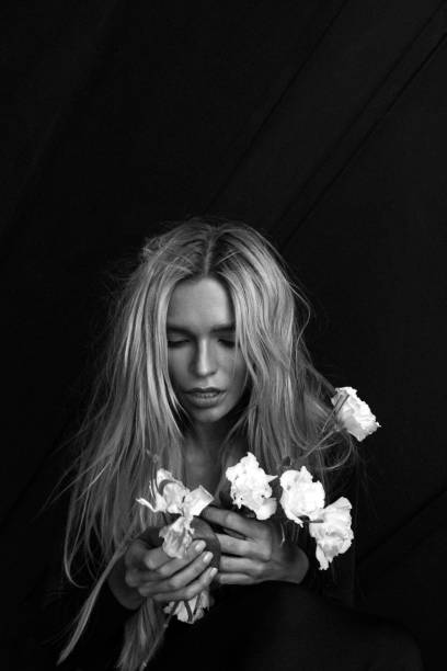 The image portrays a deeply introspective woman with loose, flowing hair, cradling delicate white flowers in her hands. Her eyes are closed as if lost in thought or emotion, encapsulating a quiet moment of solitude and reflection against a dark, minimalist background. The contrast of her expression and the soft texture of the flowers evokes a profound sense of vulnerability and grace.