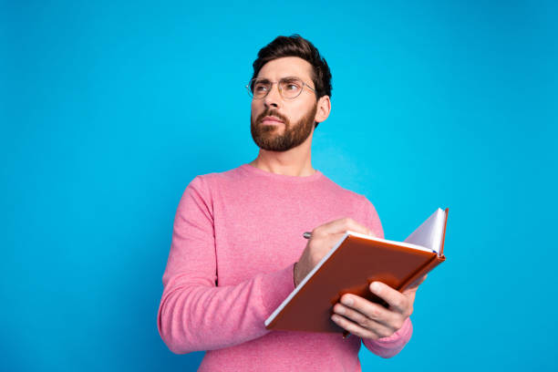 istock Confident young man in pink sweater holding notebook, thinking with determination against a blue background 2189449037