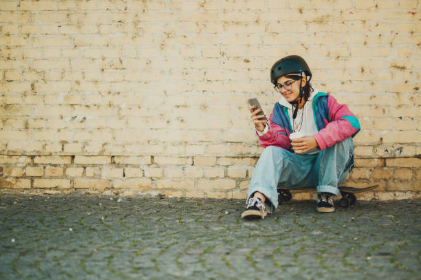 Smiling young female skater with helmet and colorful jacket is using smartphone and drinking coffee while sitting on skateboard against a brick wall