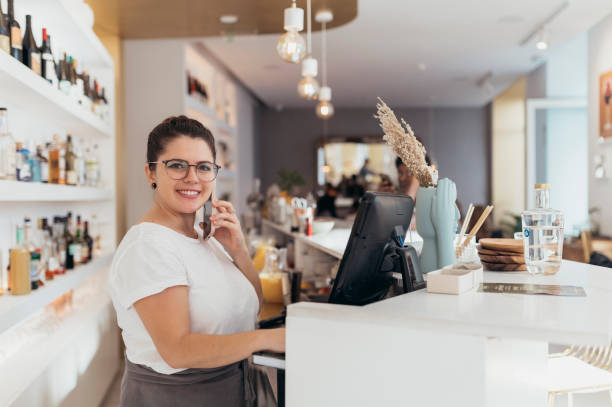 istock Waitress using cellphone at cash register, smiling at camera behind service counter in restaurant and bar 2189460010