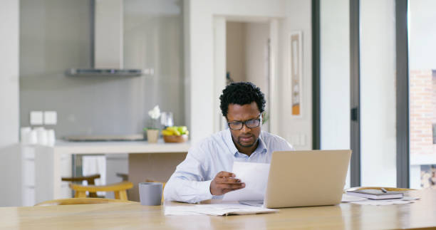 istock Businessman, laptop and documents with remote work, agency and ideas for solution or brainstorming. African, entrepreneur or brand consultant with project, proposal or online startup in home kitchen 2189468097