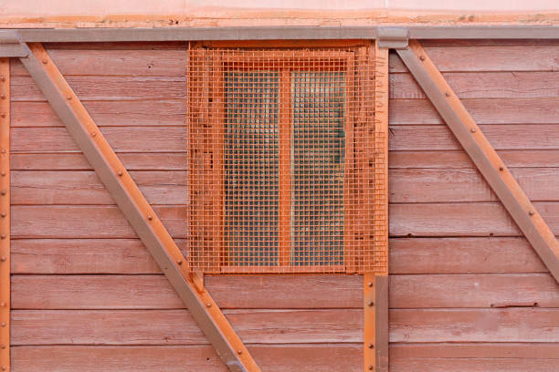 A window with a mesh screen on it is on a red wall. The window is small and the mesh screen is made of metal