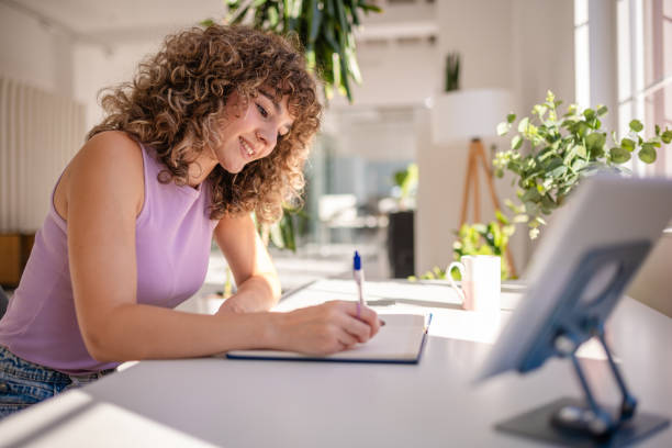 Young woman writing at desk in bright modern workspace with a tablet