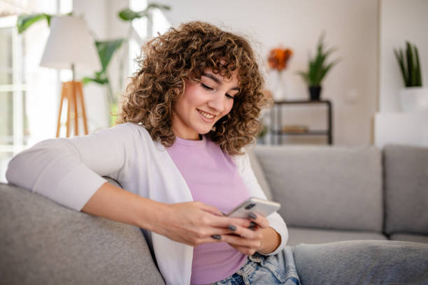 istock Young woman smiling while using a smartphone sitting on a cozy couch 2189537794
