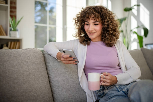 Woman relaxing at home using smartphone while holding a mug on the sofa