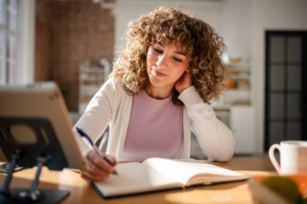istock Woman writing in a notebook while working on a tablet in a bright room 2189540215