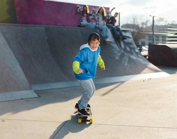 Full length of happy boy rides a skateboard while children cheer in the background during sunny day in skateboard park